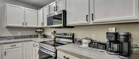 Una cocina moderna con armarios blancos, horno empotrado, microondas y una estación de café con máquina Keurig.