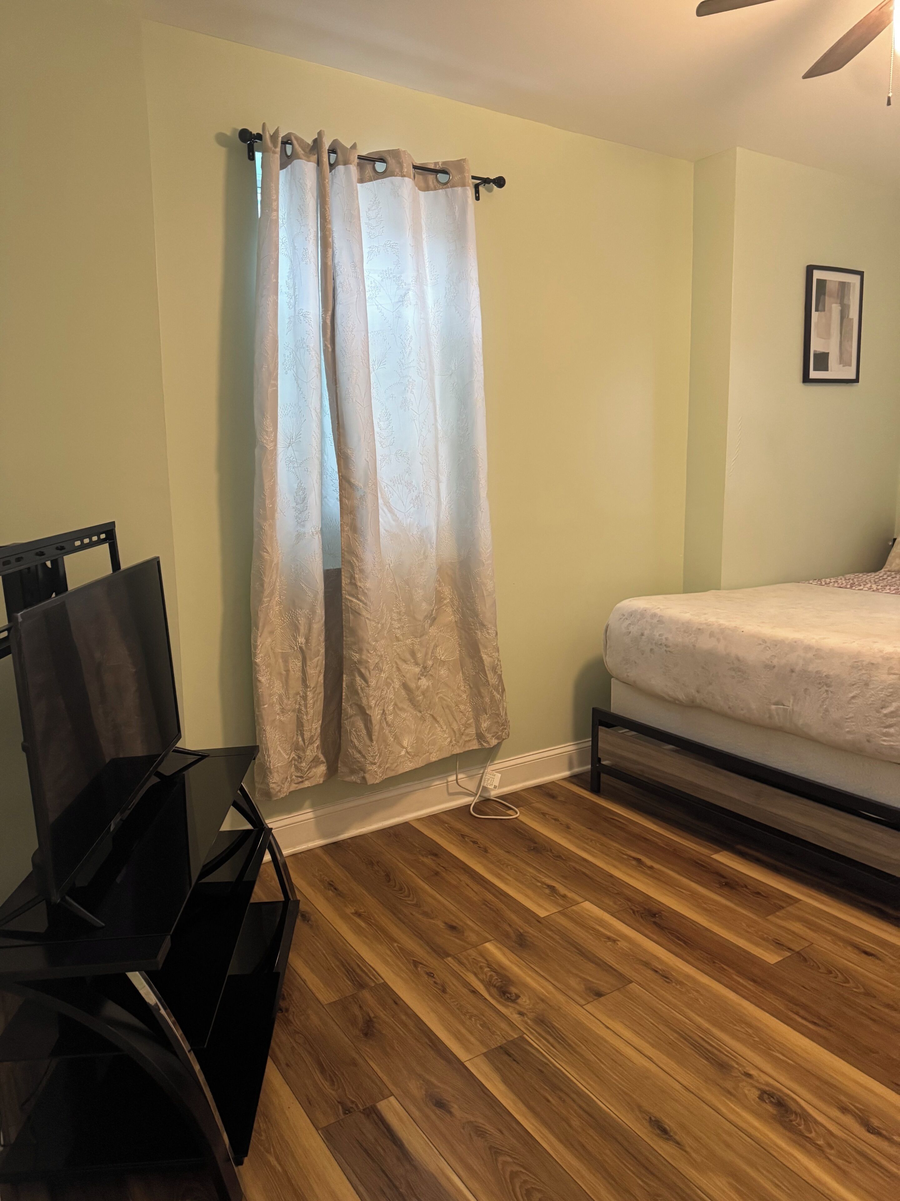 A bedroom with a bed, a wooden floor, a black desk, and a window with a curtain.