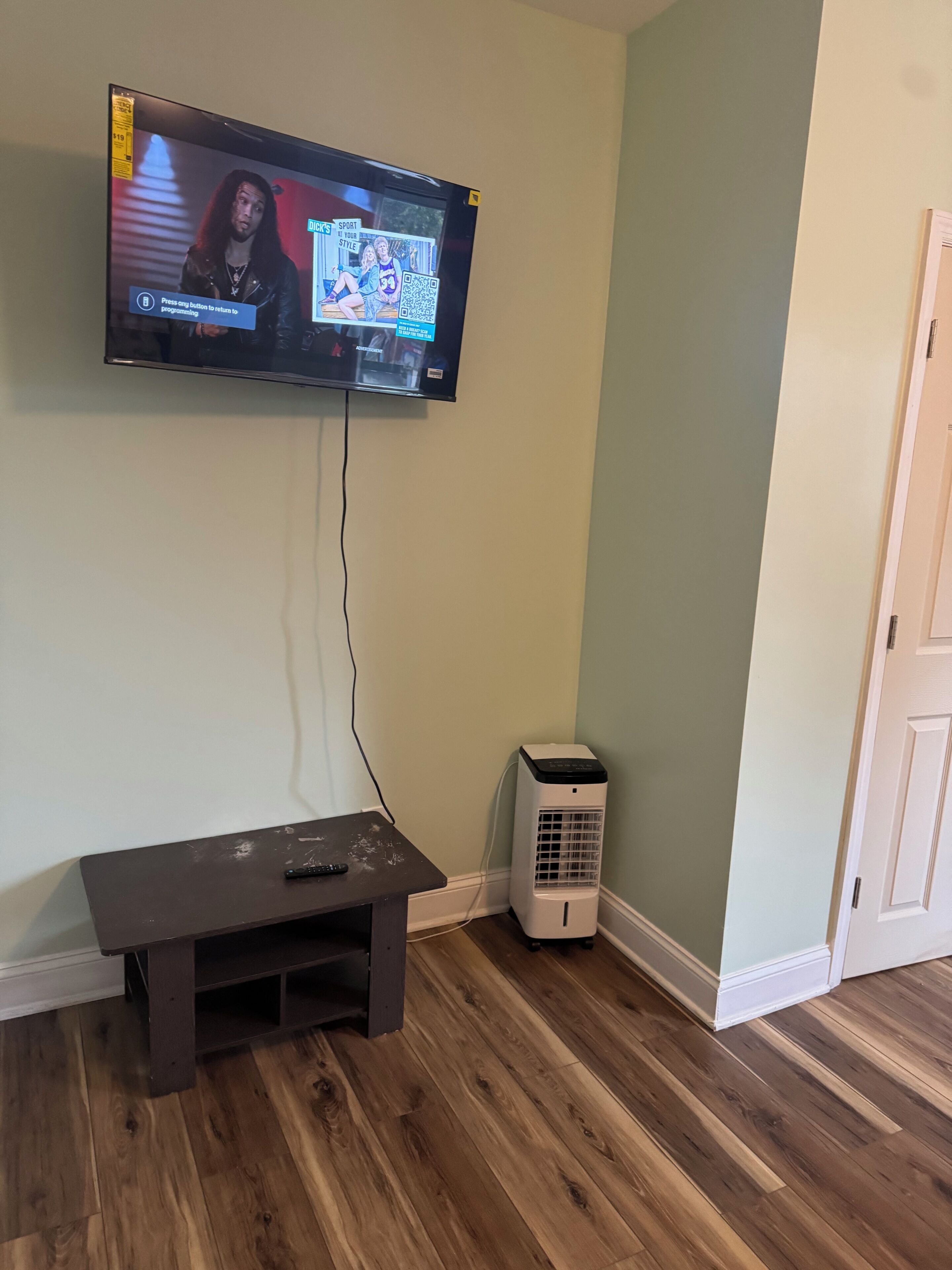 Een televisie aan een muur bevestigd, een klein tafeltje met een afstandsbediening en een witte, verplaatsbare heater.