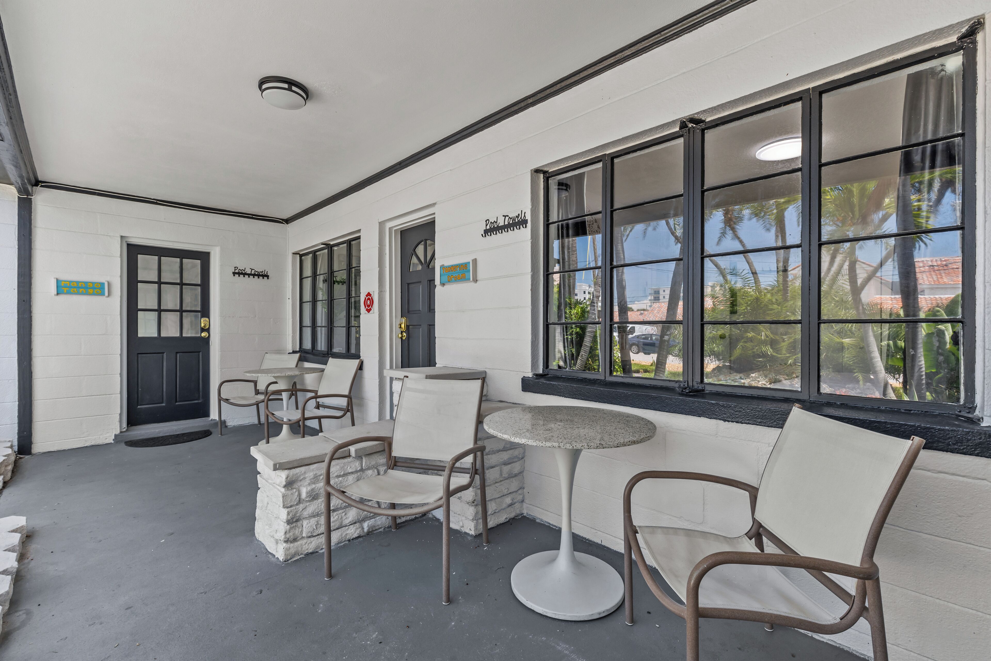A covered outdoor seating area with tables and chairs, a black door, and a window with a view of palm trees.