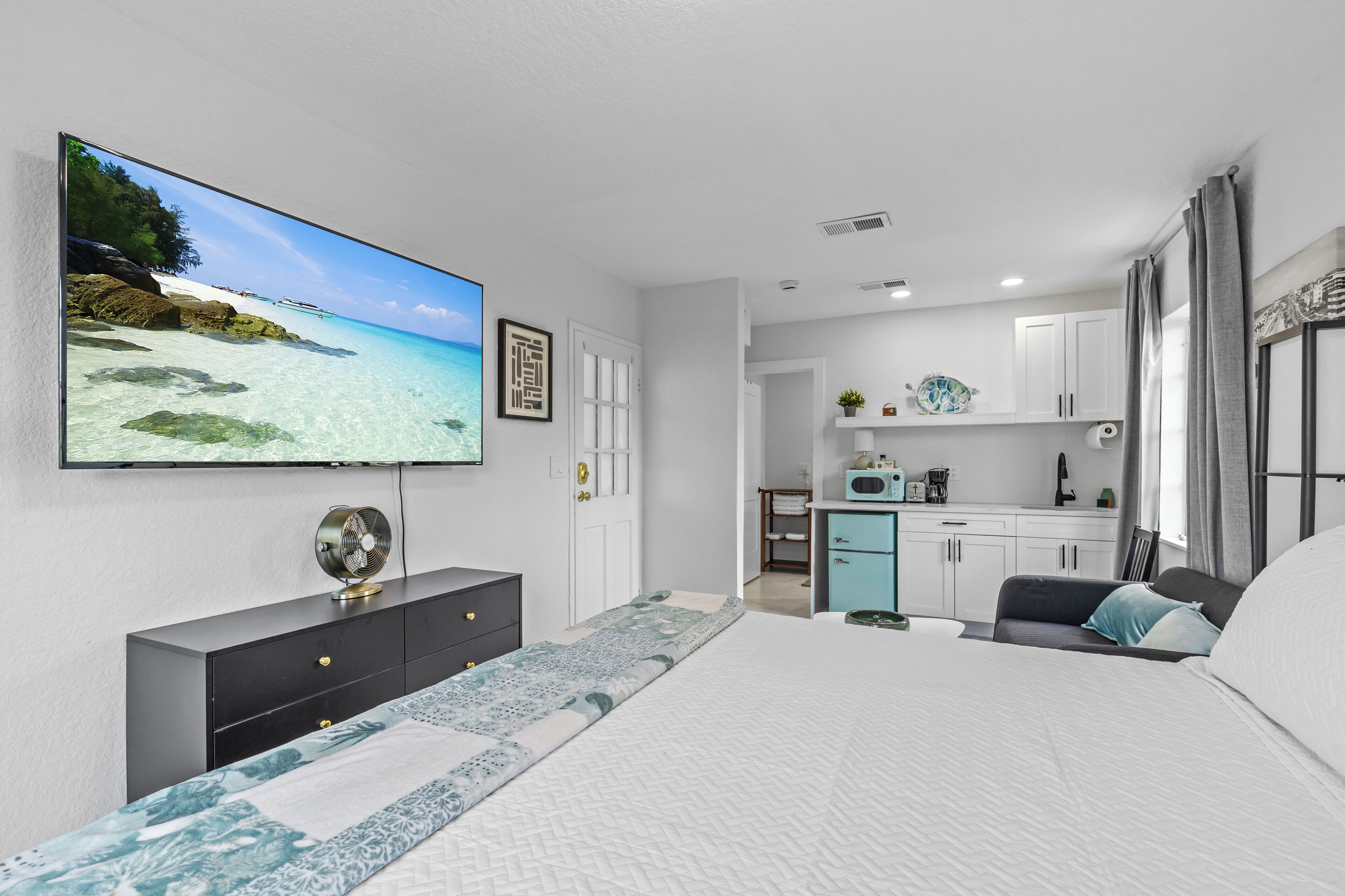 A modern bedroom with a large bed, a flat-screen TV, a dresser, and a kitchenette in the background.
