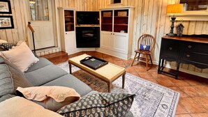 Ein gemütliches Wohnzimmer mit einer Couch, einem Couchtisch und einem an einer Holzwand montierten Fernseher. Im Raum befindet sich außerdem ein Bücherregal und ein Sessel.