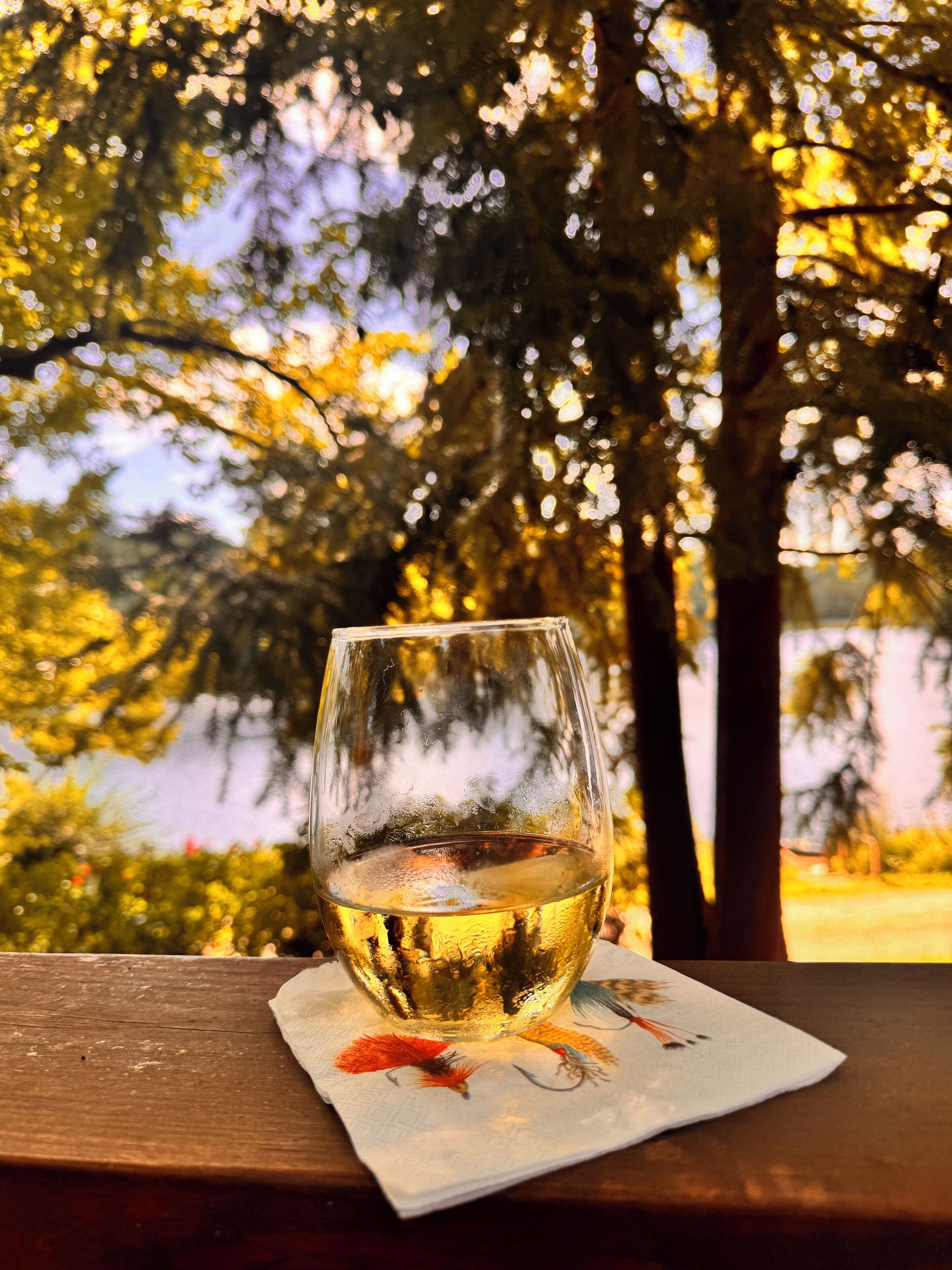 A glass of white wine on a wooden table with a napkin underneath, set against a backdrop of trees and a body of water.
