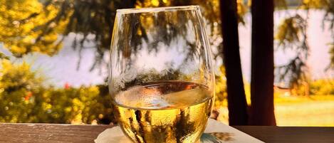 A glass of white wine on a wooden table with a napkin underneath, set against a backdrop of trees and a body of water.