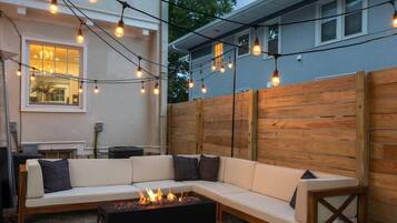 Un espacio al aire libre acogedor con un sofá modular, una chimenea y luces colgantes.