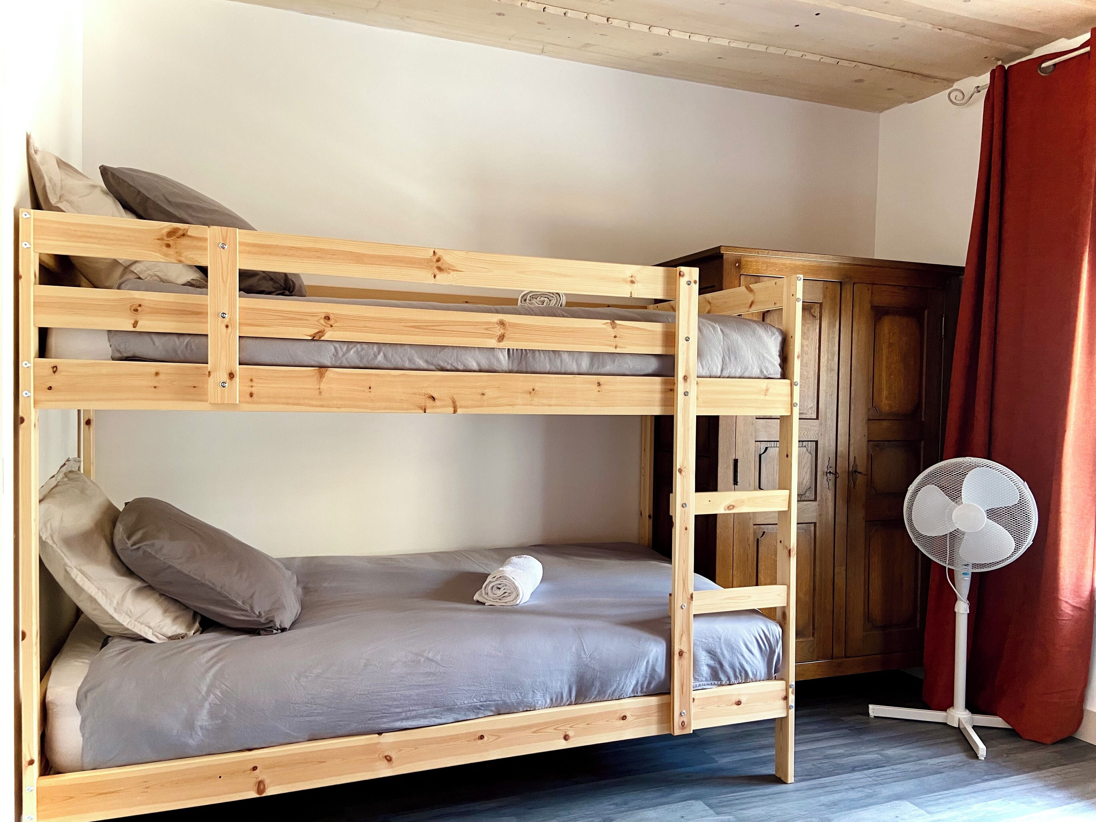 A bunk bed with a wooden frame, grey bedding, and a rolled towel on the lower bunk. A white fan is on the floor beside the bed.