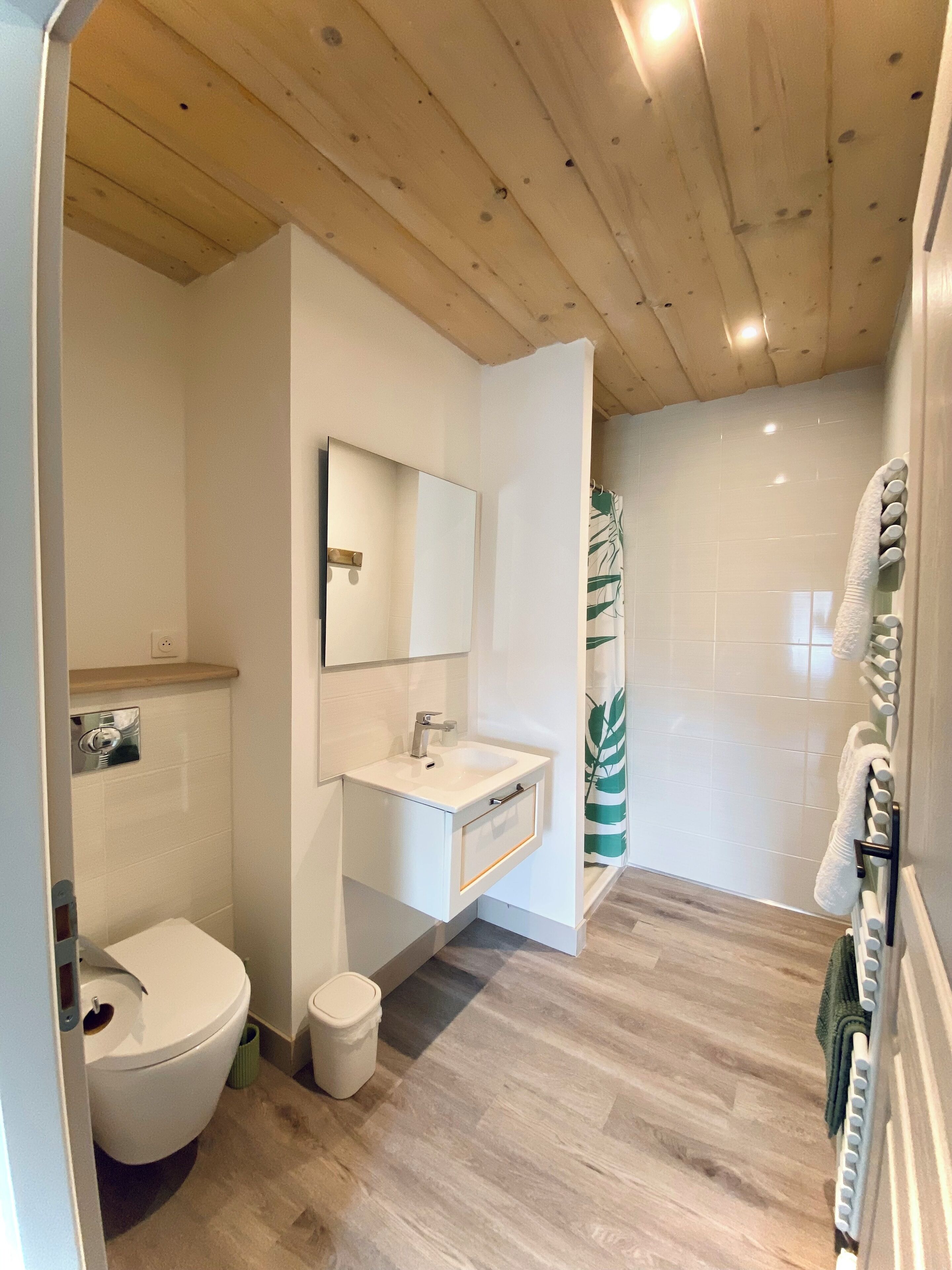 A modern bathroom with a wooden ceiling, white tiles, a sink with a mirror, a toilet, and a shower area with a towel rack.
