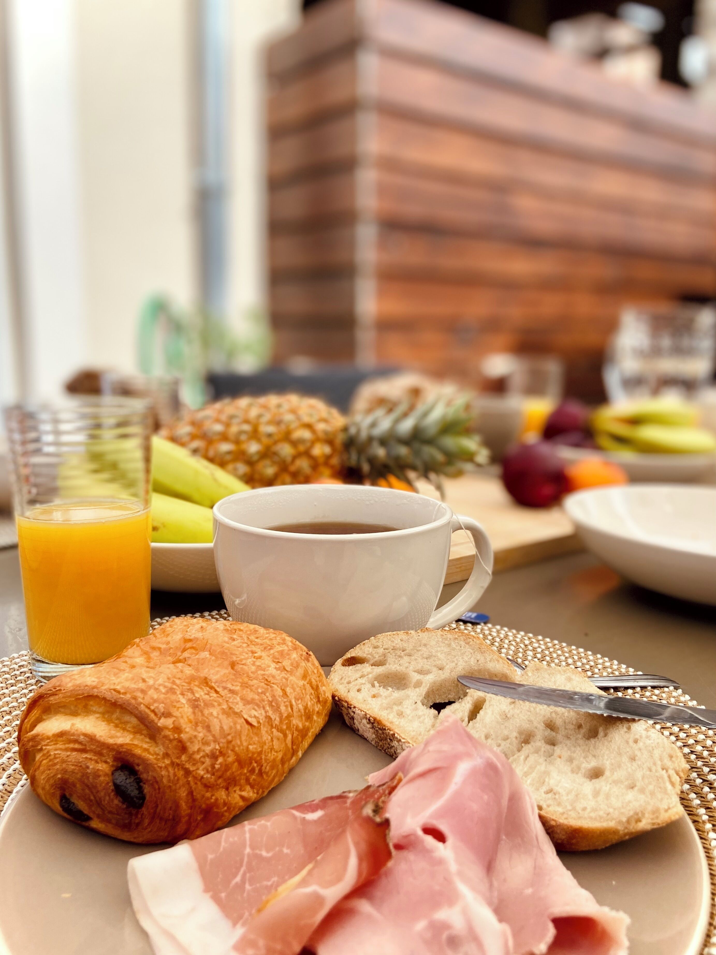 A breakfast setting with croissants, ham, bread, a pineapple, and a glass of orange juice.