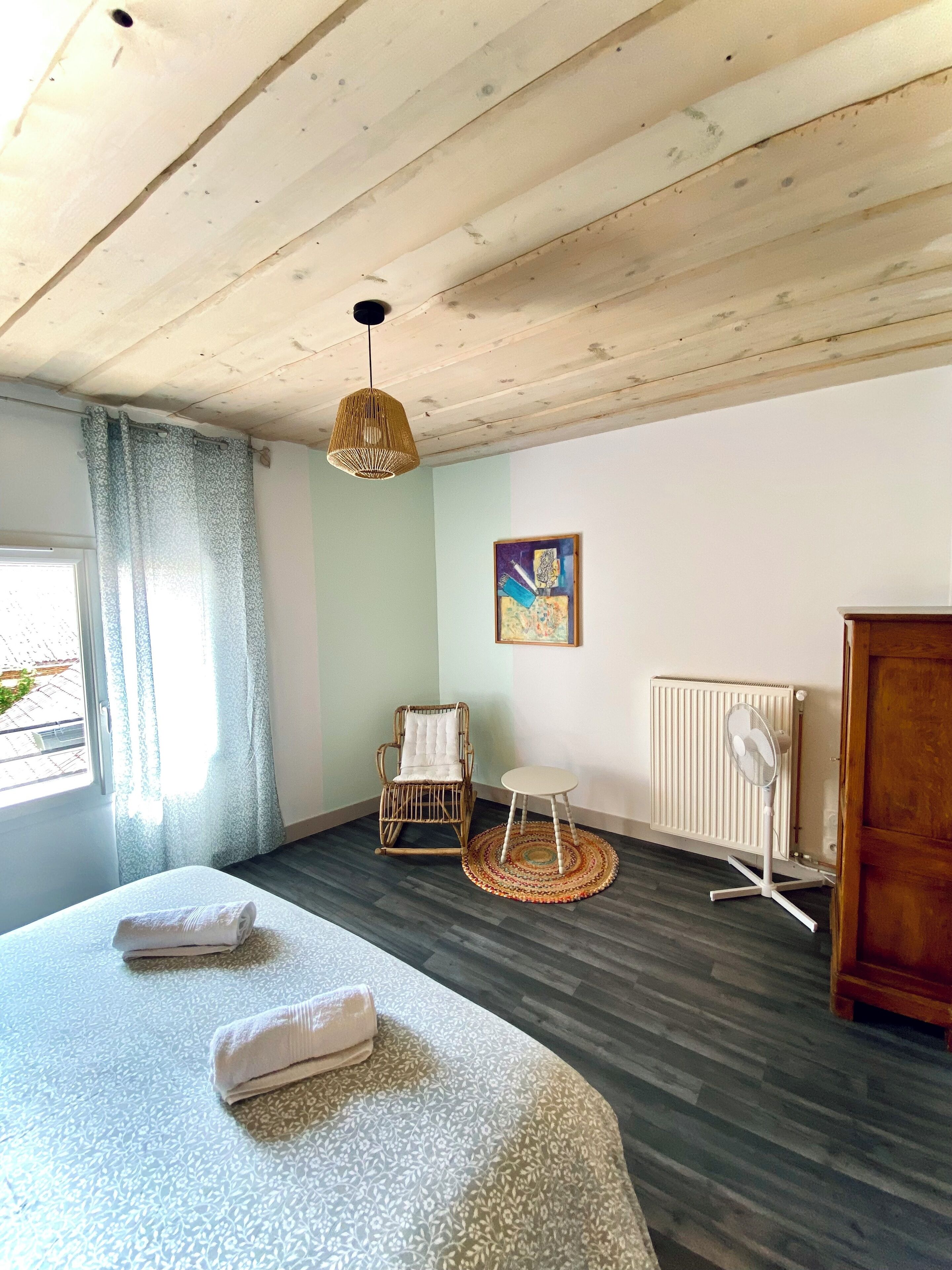 A room with a large bed, a wicker chair, a small table, a radiator, and a painting on the wall.