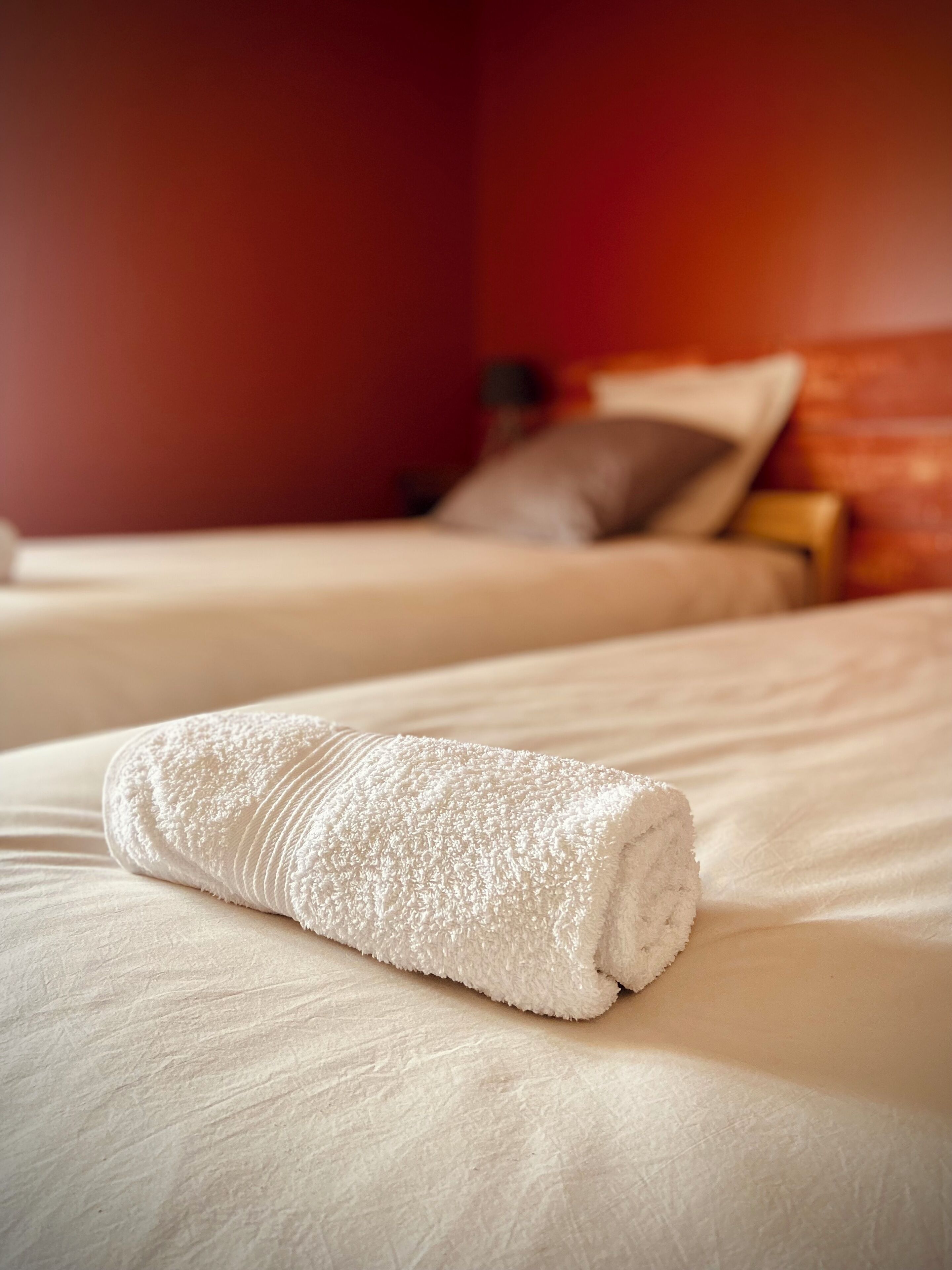 Un lit bien fait, avec une serviette blanche roulée, dans une chambre d’hôtel aux murs rouges et au chevet en bois.