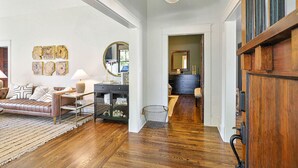 Un salón amplio con sofá de cuero, una mesa de centro y una alfombra. Se observa un pasillo que conduce a un dormitorio con puerta de madera.