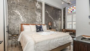 Un dormitorio con una cama grande, cabecera de madera, un taburete pequeño, una mesita de noche, una lámpara y una ventana con persianas.