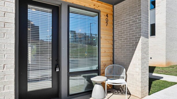 Een modern gebouw met een zwarte deur, grote ramen en een klein buitenterrein met een stoel en tafel.