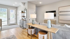 Een moderne woonkamer met een houten eettafel, een grijze bank en een klein keukenhoekje met een koelkast en een spiegel.