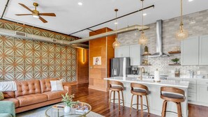 A modern living room with a sofa, coffee table, and bar stools. A kitchen with white cabinets and a stainless steel range hood is visible.