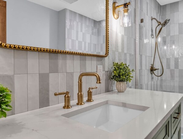 A bathroom with a white sink, gold faucets, a mirror with a gold frame, and a shower with a gold showerhead.