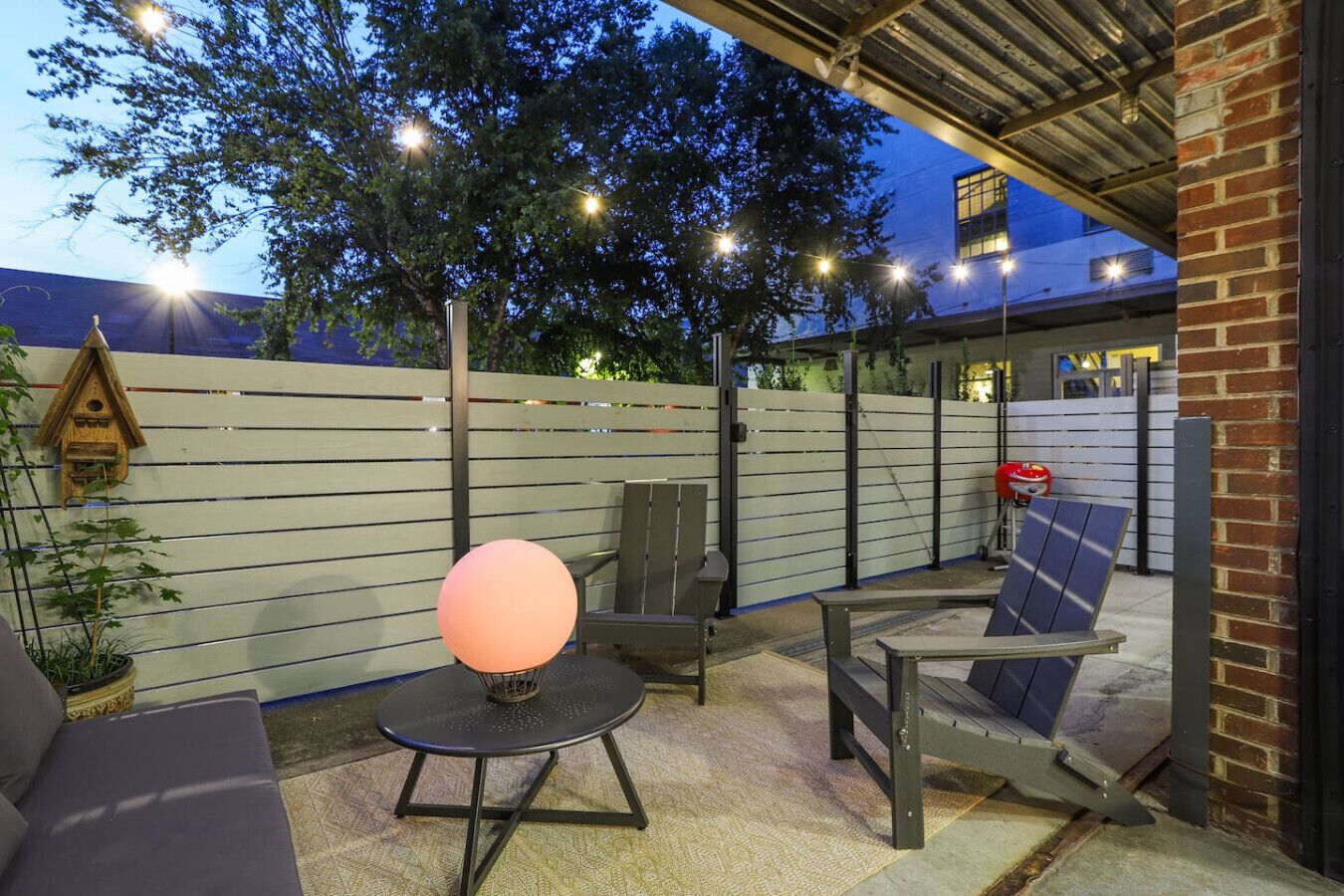 An outdoor patio area with a round table, a chair, and a sofa. There is a decorative light fixture on the table and a birdhouse on the wall.