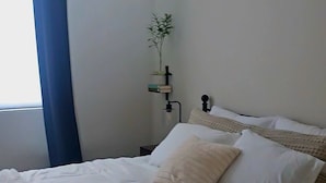 A neatly made bed with white bedding and two pillows, a bedside table with a lamp, and a window with blue curtains. - Pleasant 3-bedroom house with AC in San Tan Valley (San Tan Valley)