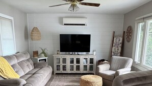 Un salon avec un canapé gris, un fauteuil gris, un téléviseur posé sur un meuble blanc, un ventilateur de plafond et une fenêtre donnant sur un paysage verdoyant.
