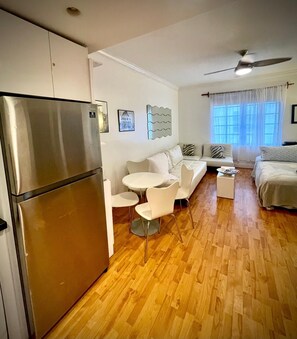 A modern, well-lit room with a wooden floor, a stainless steel refrigerator, a white sofa, a small round table, and a bed with a white duvet. - Awesome Location Heart of South Beach -Walk to Beach, Shops & Nightlife (Miami Beach)