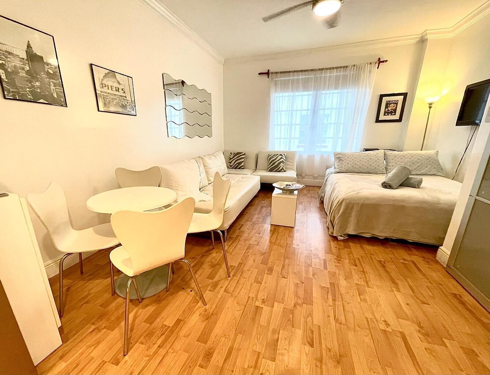 Ein modernes, helles Zimmer mit einer Couch, einem Bett, einer Essecke mit Stühlen und einem kleinen Tisch. An der Wand hängen Bilderrahmen und ein Fenster mit Vorhängen.