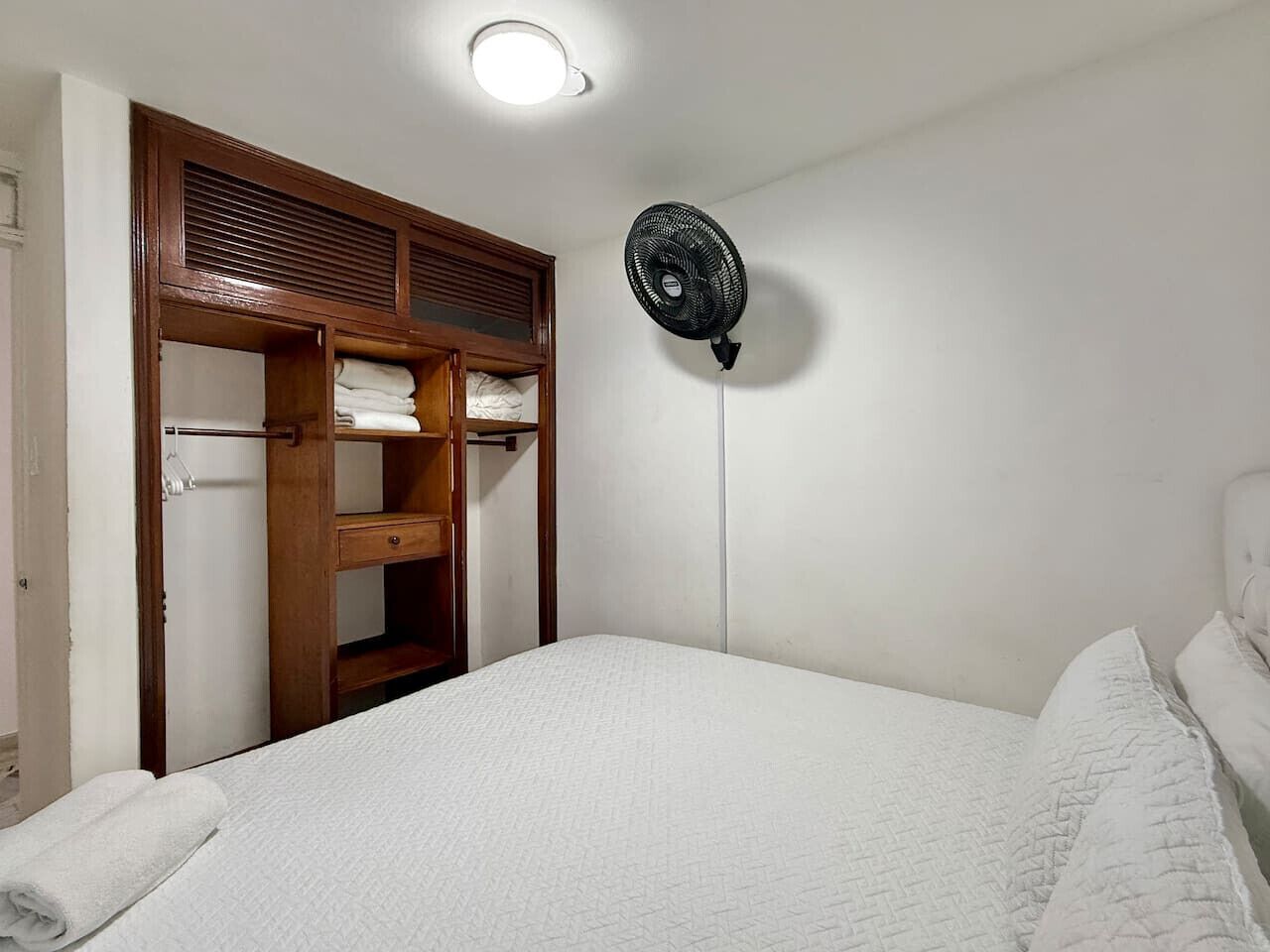 A bedroom with a bed, a wooden wardrobe, and a wall-mounted fan.