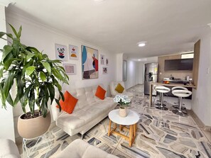 A modern living room with a sofa, a coffee table, and a potted plant. There is a kitchen area with a refrigerator and a sink.