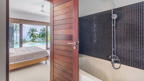A modern bathroom with a bathtub and shower, a wooden door, and a view of a pool and palm trees. - Serene Bay View Villa in Ko Samui - Your Tropical Paradise (Koh Samui)
