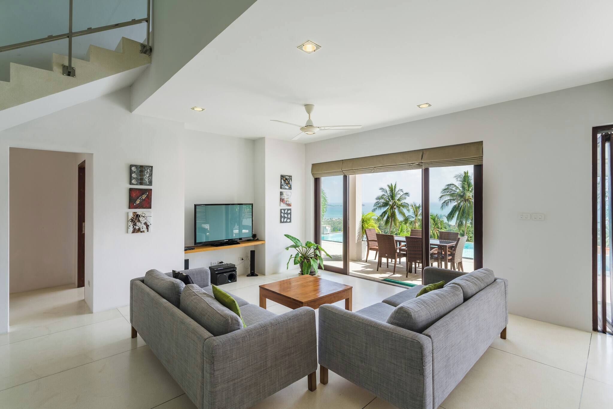 A modern living room with grey sofas, a wooden coffee table, a flat-screen TV, and a large sliding glass door leading to a balcony with outdoor furniture.
