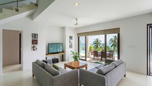 A modern living room with grey sofas, a wooden coffee table, a flat-screen TV, and a large sliding glass door leading to a balcony with outdoor furniture. - Serene Bay View Villa in Ko Samui - Your Tropical Paradise (Koh Samui)