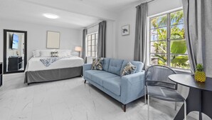 Una habitación de hotel moderna con cama, sofá, mesa de comedor y un gran ventanal con cortinas.