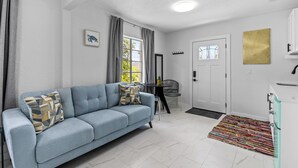 Una sala moderna con un sofá azul, un comedor con una mesa pequeña y sillas, una cocina con armarios blancos, y una alfombra estampada en el suelo.