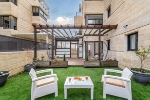 A courtyard with a pergola, two white wicker chairs, a table with drinks, and planters with small trees and flowers. - Luxury 5BR Huge Oasis by the Shuk (Jerusalem)