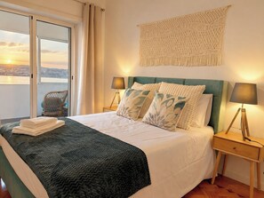 A hotel room with a bed, two bedside lamps, a view of the city through a window, and a balcony. - Flat in Cacilhas – Lisbon Stunning River Views (Almada)
