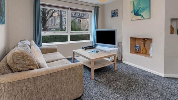 A living room with a sofa, a TV on a stand, a small table, a window with curtains, and a wall-mounted shelf with decorative items.