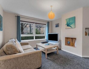 A living room with a sofa, a TV on a stand, a small table, a window with curtains, and a wall-mounted shelf with decorative items. - Charming Edinburgh Apartment With City Views (Edinburgh)