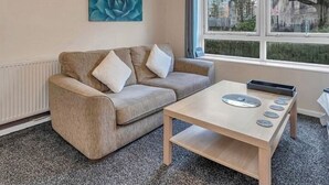 A room with a beige sofa, a wooden coffee table with circular objects, a blue artwork, and a window with a view of trees and buildings.