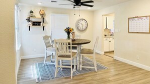 A dining area with a round table, four chairs, a ceiling fan, a wall clock, a calendar, and a kitchen in the background. - Clean, Cute, and Comfortable (Safford)