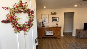 A hallway with a wooden floor, a white door with a wreath, a cabinet with a 'good tidings joy' sign, and a 'hello' sign on the wall. - Clean, Cute, and Comfortable (Safford)