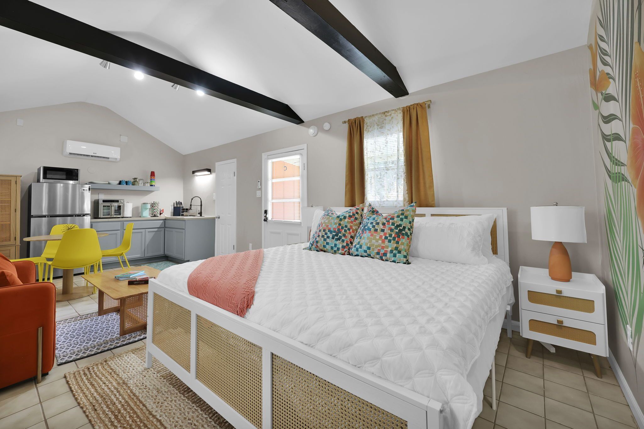 A bedroom with a bed, bedside tables, a nightstand, a chair, a desk, and a kitchen area with appliances.