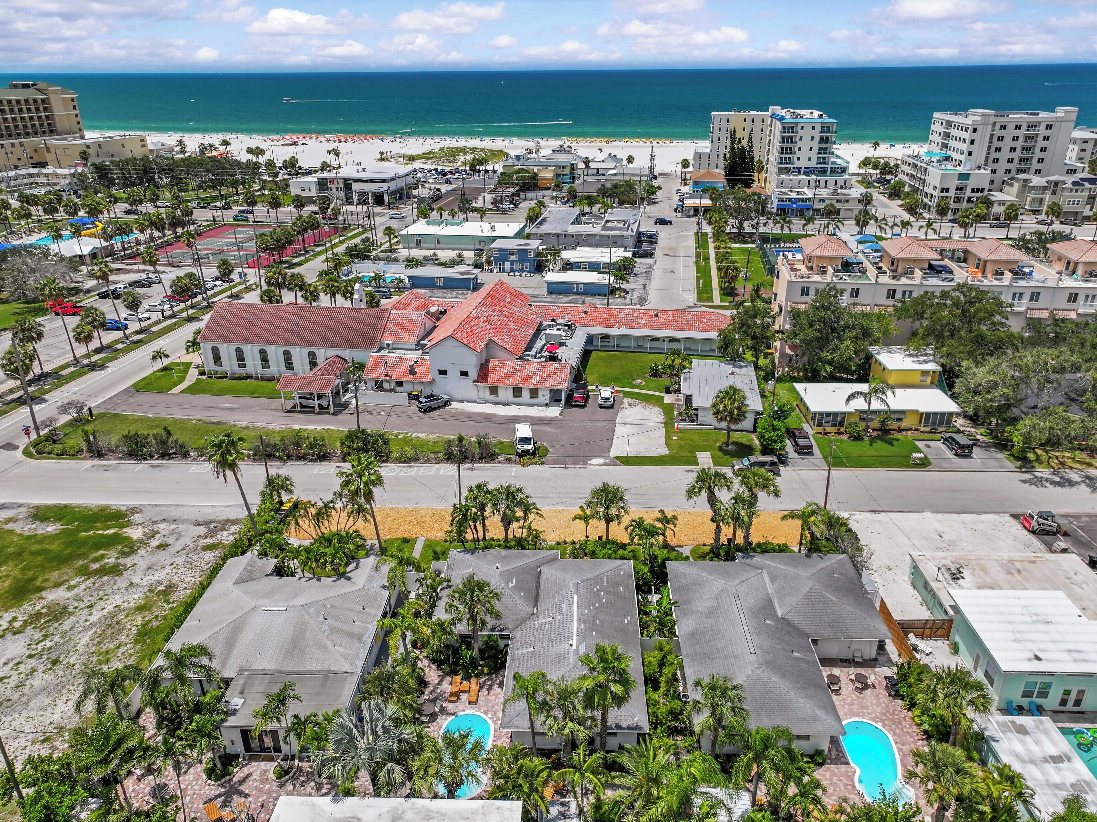 A coastal resort area with a large building featuring red-tiled roofs, a swimming pool, and a beach in the background.