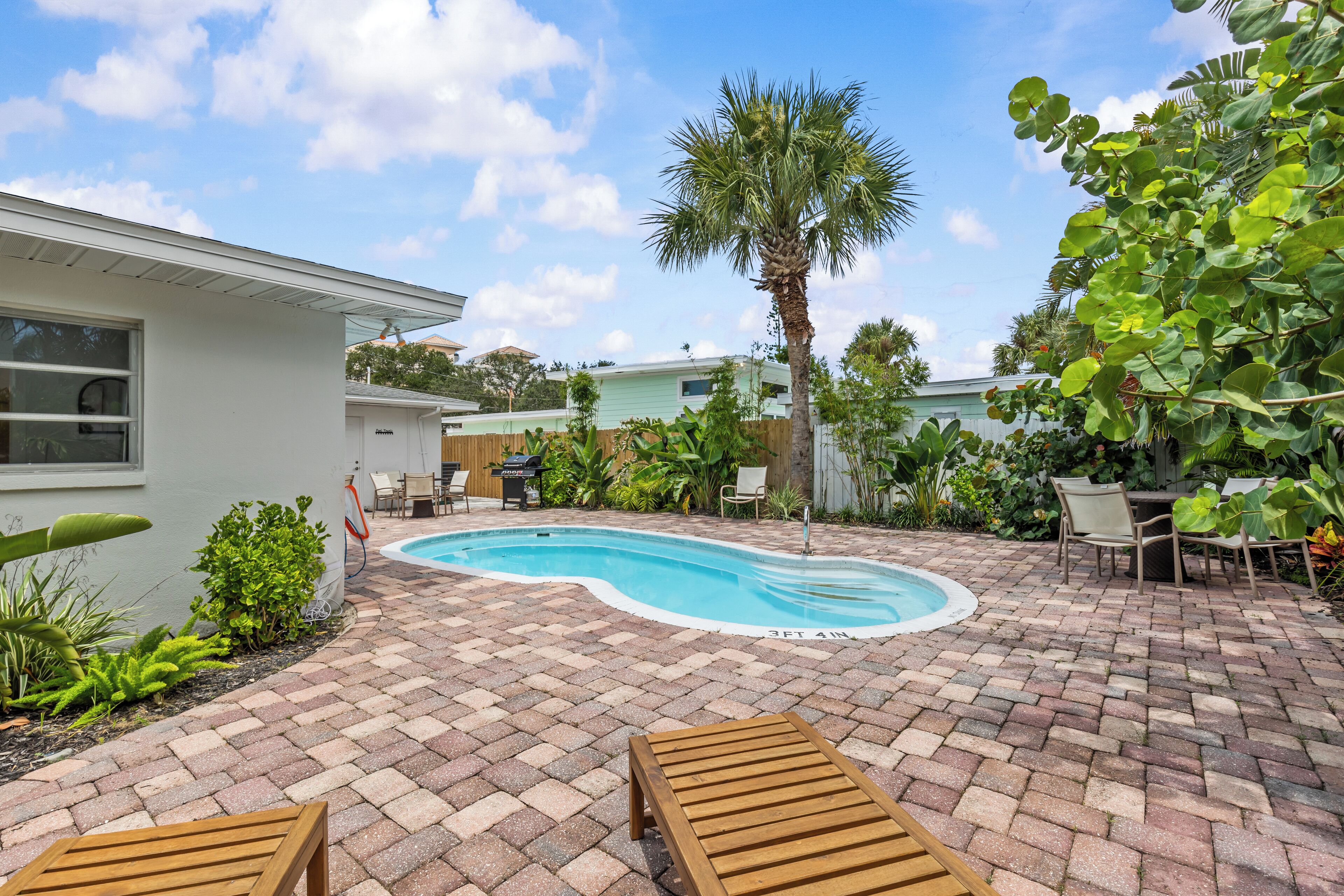 A swimming pool with a 3ft 4in depth marker, surrounded by a brick patio, wooden lounge chairs, and various plants.