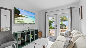A living room with a large TV, a green console, a white sofa, and a view of a beach through the sliding glass door.