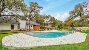 A swimming pool with a sandy deck, surrounded by trees and a grassy area. - Spacious 6-bedroom house in delightful Grand Prairie with Pool (Grand Prairie)