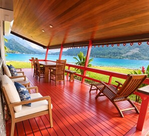 A wooden deck with a red railing, outdoor furniture, and a view of a beach and ocean.