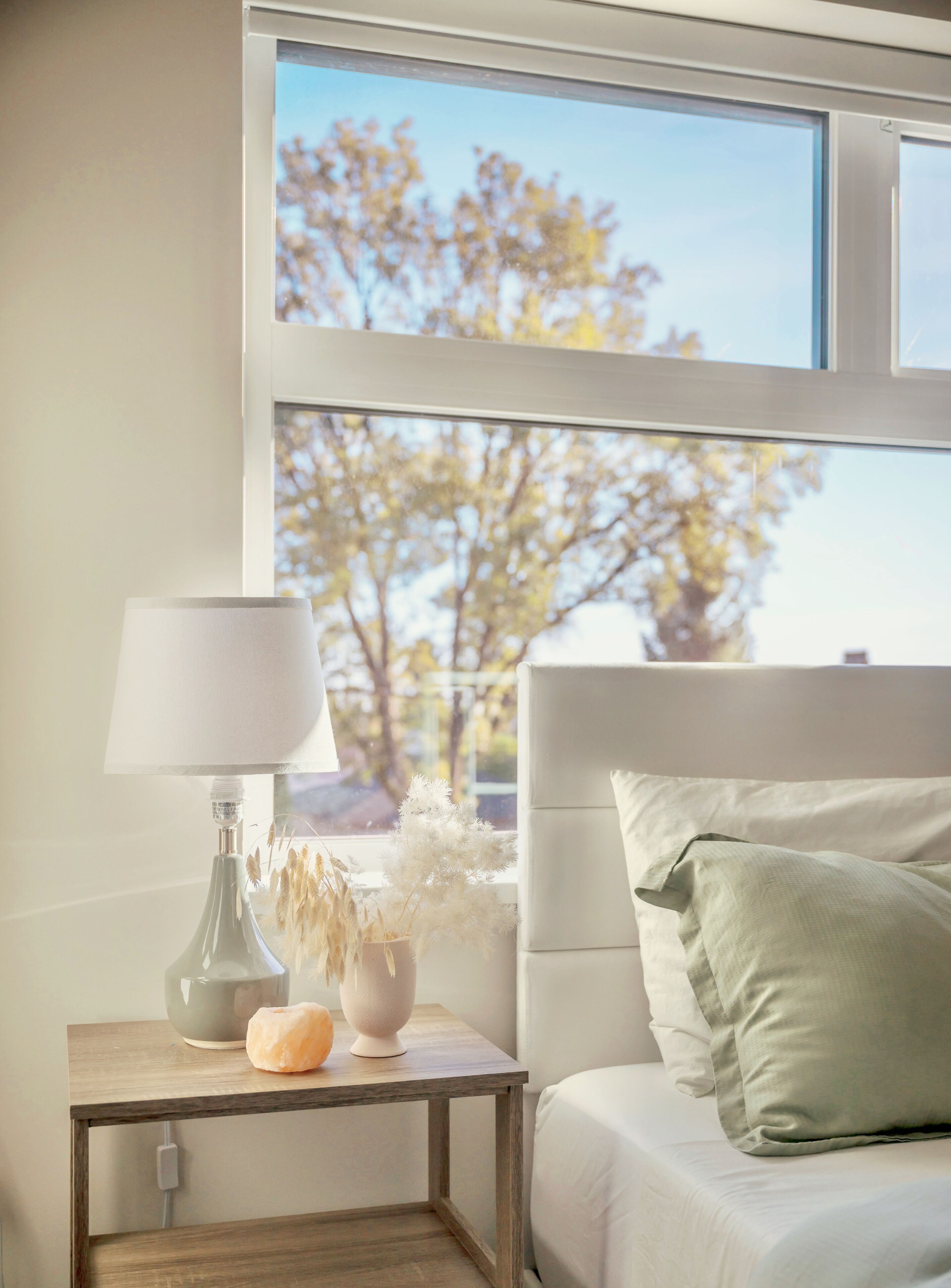 A bedroom with a bed, a bedside table with a lamp, a vase with dried flowers, and a view of trees through a window.