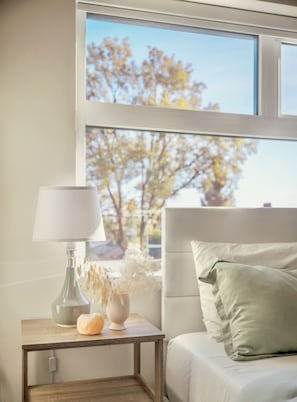 A bedroom with a bed, a bedside table with a lamp, a vase with dried flowers, and a view of trees through a window.