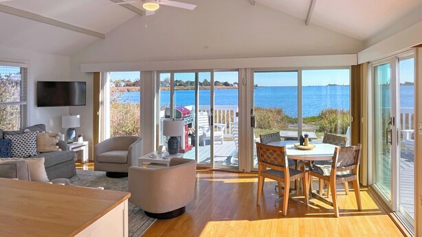 Een ruime woonkamer met een grote televisie, comfortabele zitmogelijkheden en uitzicht op de zee door grote glazen deuren.