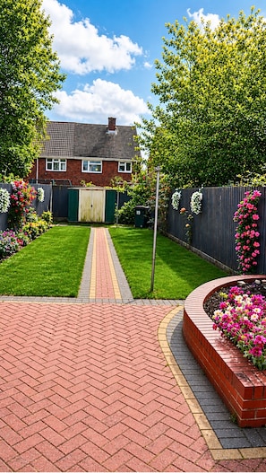 A residential backyard with a paved pathway, a brick wall, and a house with a visible garage door.