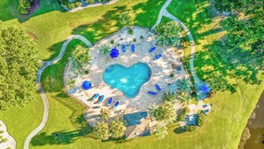 Vista aérea de una piscina con forma de corazón rodeada de sillones de descanso y sombrillas, con senderos y vegetación.